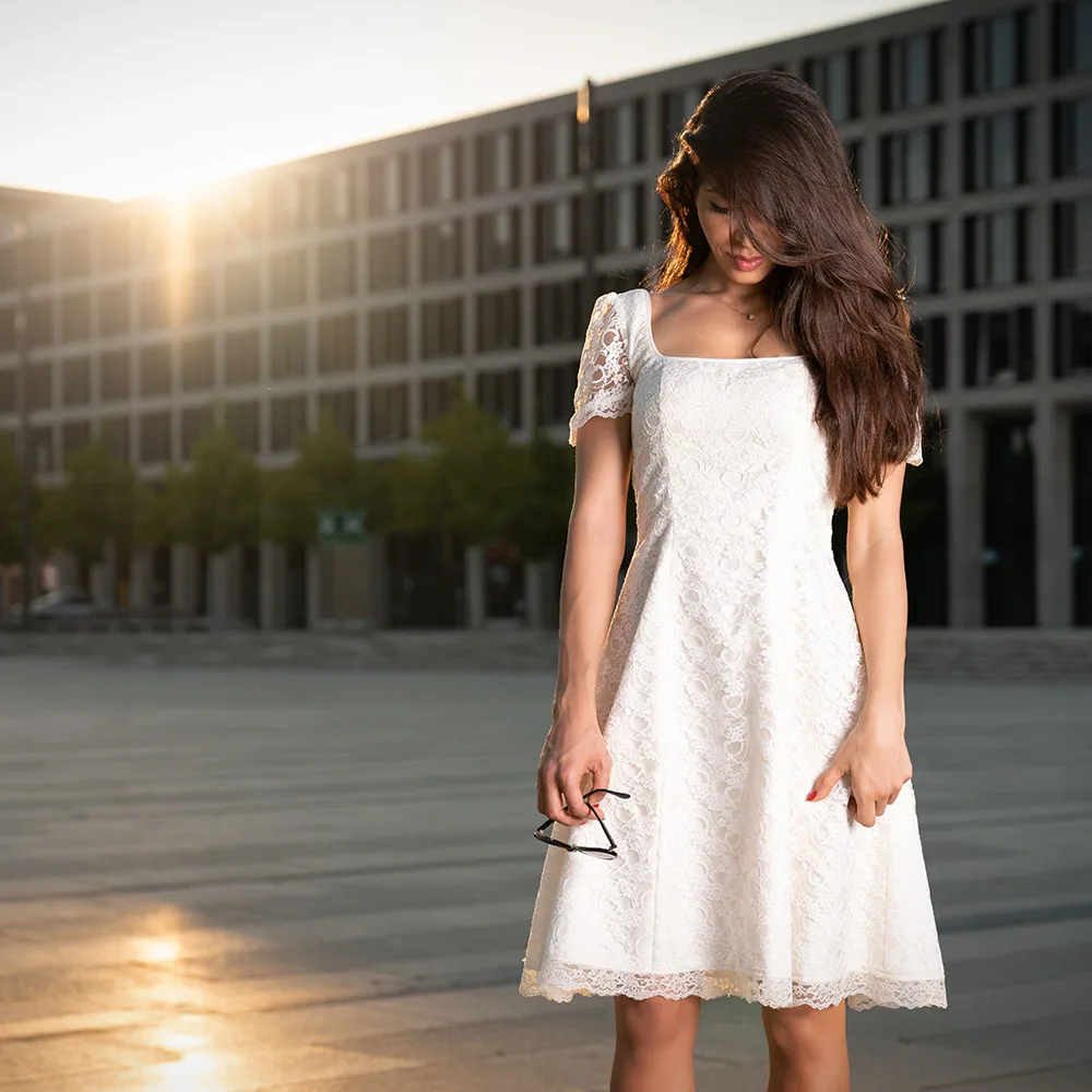 A woman in a white lace dress in front of a modern building at sunset