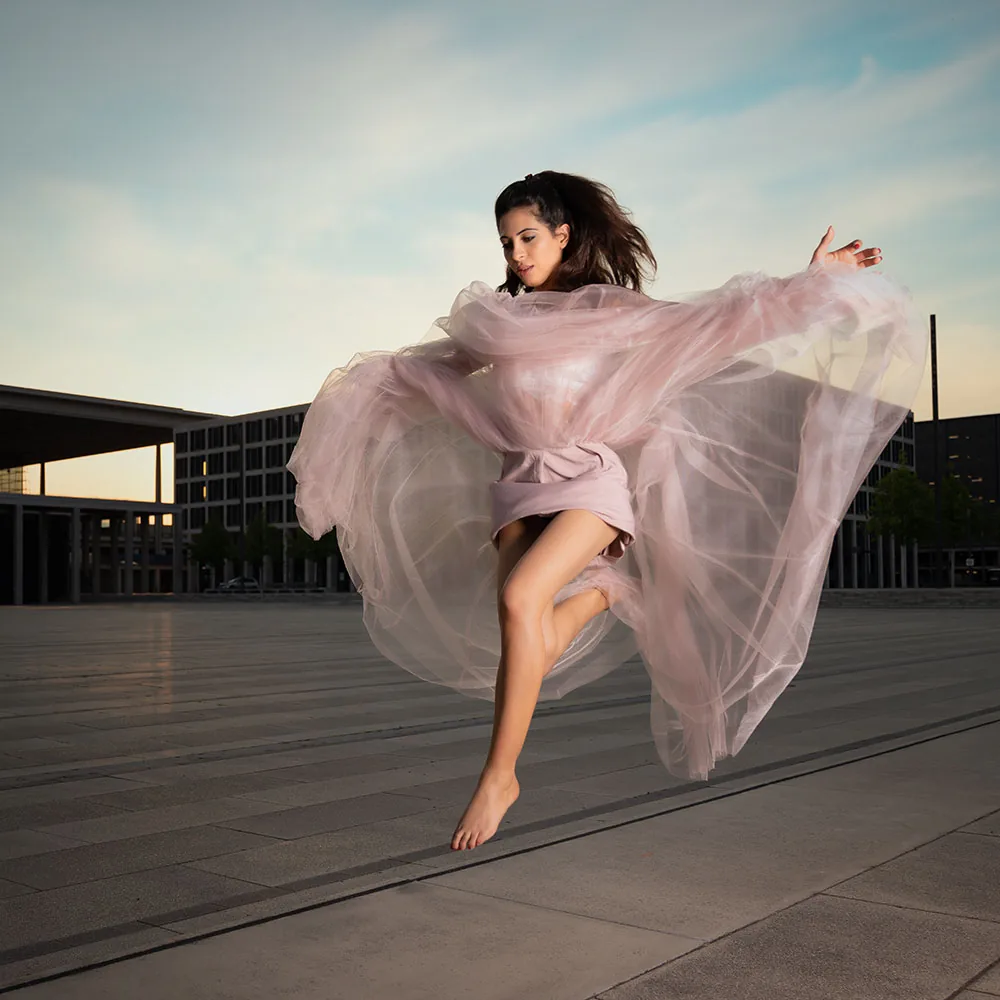 A dancer in a flowing pink dress leaps across an empty square at dusk